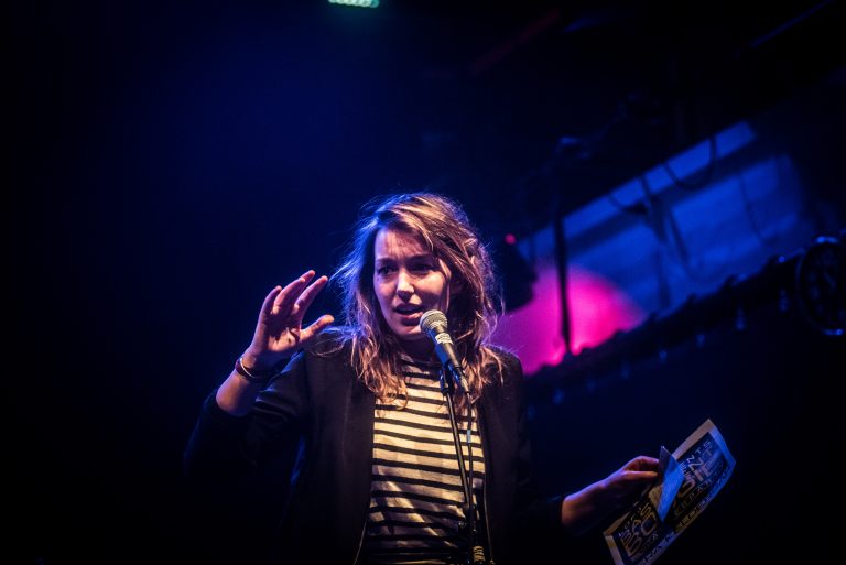 Een vrouw met een microfoon staat op een podium onder kleurrijke verlichting en spreekt met haar hand opgestoken.