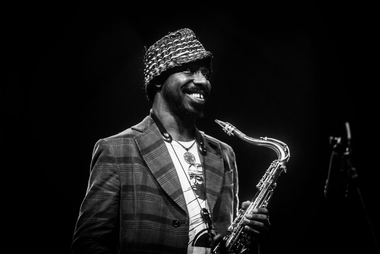 Een muzikant met een saxofon staat glimlachend op het podium, gekleed in een gestreepte blazer en een opvallende hoed.