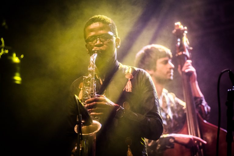 Een muzikant speelt saxofon op een podium met sfeervolle belichting, terwijl een andere muzikant op de achtergrond zichtbaar is.