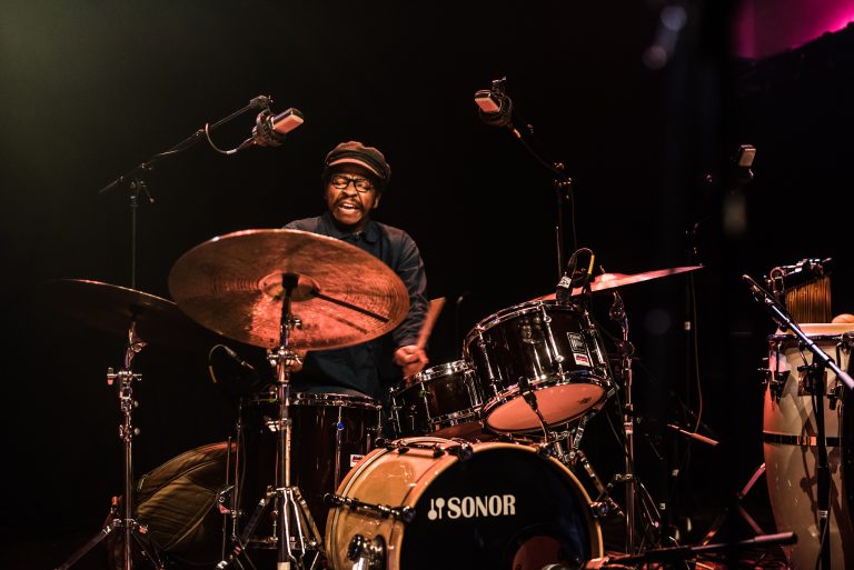 Een drummer speelt energiek op zijn drumstel met een glimlach, omgeven door microfoons en een donkere achtergrond.