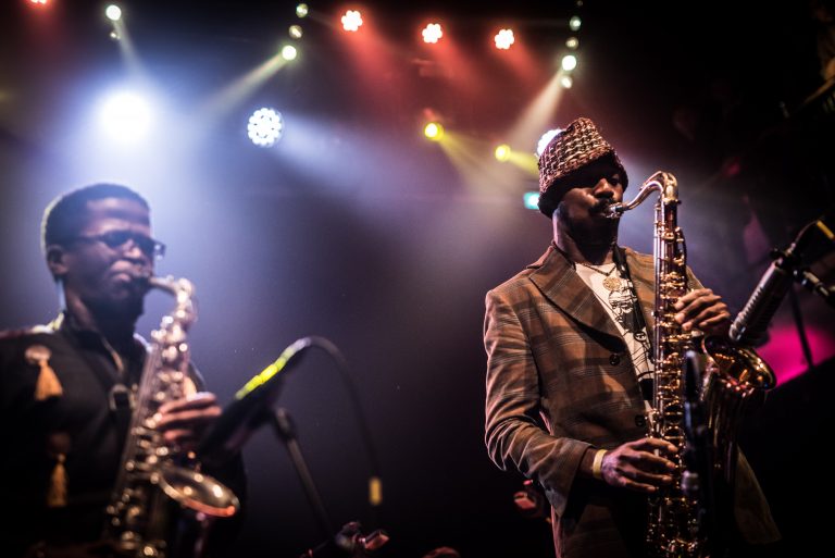 Twee saxofoonspelers op het podium, omringd door kleurrijke lichten tijdens een liveoptreden.