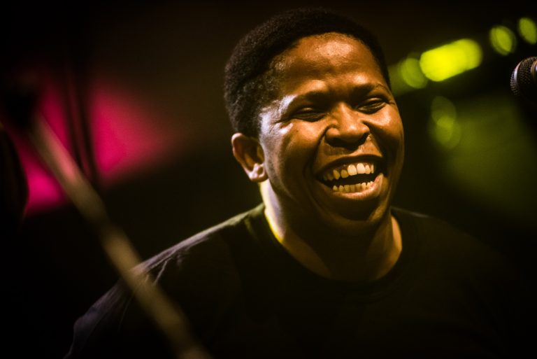 Een glimlachende man met een donkere huid staat in een kleurrijke, sfeervolle omgeving.