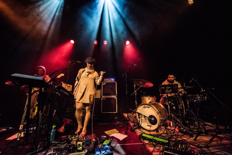 Een band speelt live op het podium met kleurrijke lichtstralen en muzikanten die verschillende instrumenten bespelen.