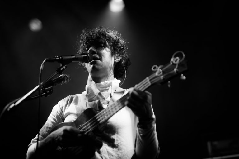 Een muzikant met een krullend kapsel staat op het podium en speelt een basgitaar terwijl hij zingt.