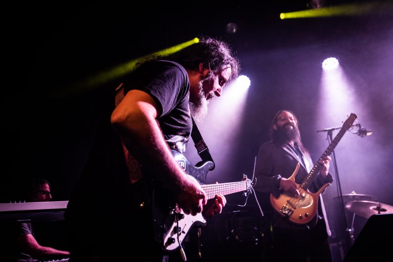 Twee muzikanten spelen gitaar op een podium, omringd door sfeervolle verlichting.