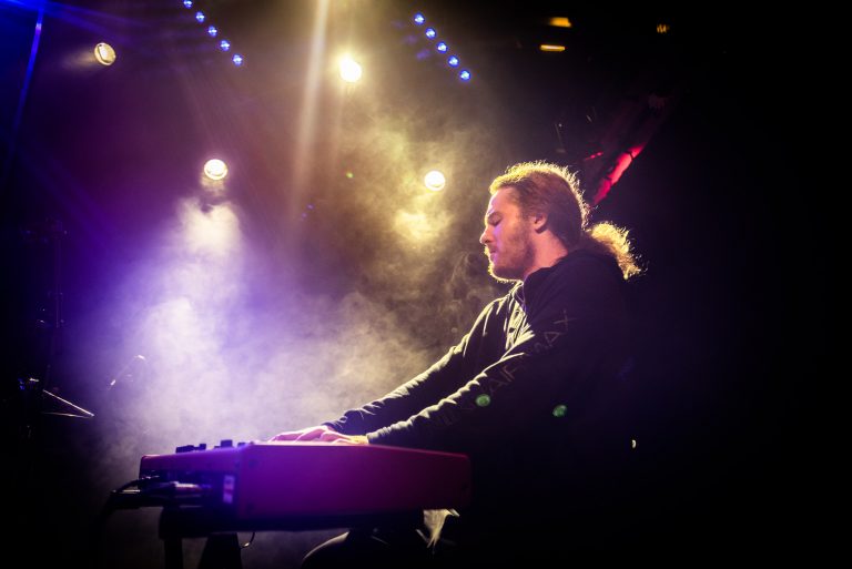 Een muzikant speelt op een keyboard terwijl kleurrijke lichten en rook om hem heen draaien.