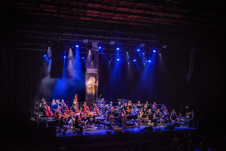 Een groot orkest speelt op een podium met blauwe verlichting.