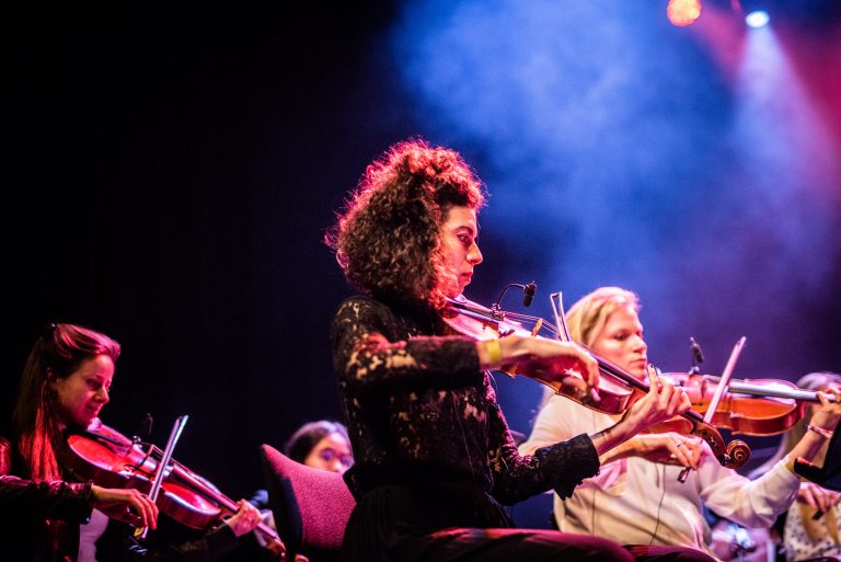 Een vrouw speelt viool op het podium, omringd door andere muzikanten, met kleurrijke verlichting en een rookachtige achtergrond.