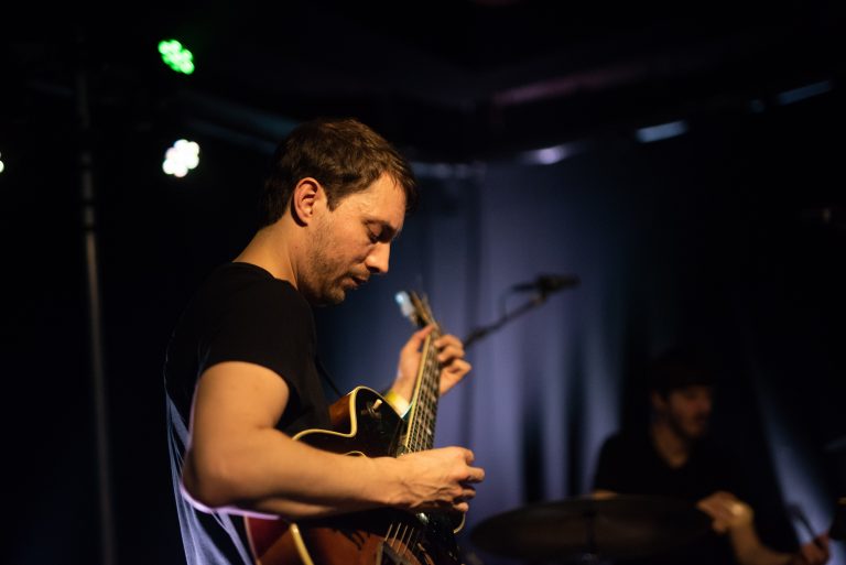 Een muzikant speelt gitaar op een podium met een donkere achtergrond en sfeerverlichting.