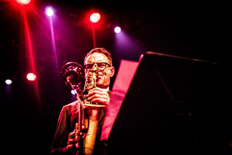 Een muzikant speelt saxofon op een podium met kleurrijke verlichting.