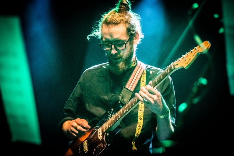 Man met een baard en een knotje speelt gitaar op een podium met kleurrijke verlichting.