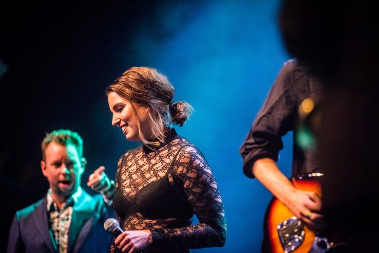 Een zangeres in een zwart kledingstuk zingt op een podium, omgeven door bandleden.