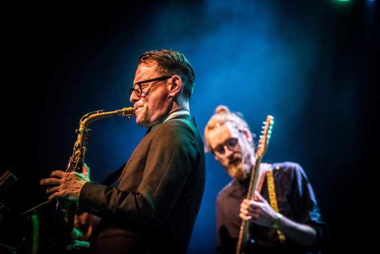 Een saxofonist speelt gerichter in een live muzieksetting, terwijl een gitarist op de achtergrond toekijkt.