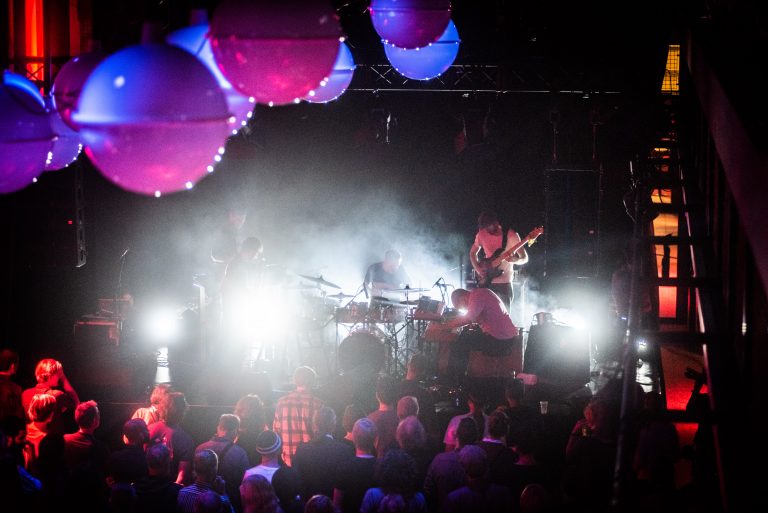 Een muzikant performt op een podium voor een levendig publiek, omringd door kleurrijke verlichting en rook.