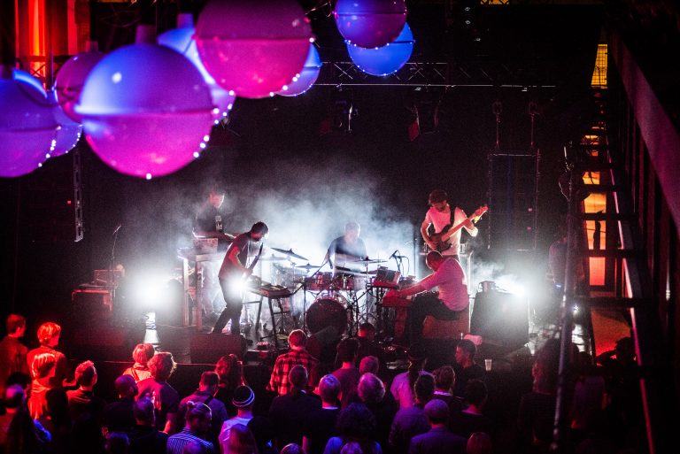 Een liveband speelt op een podium, omringd door kleurrijke lichten en een menigte toeschouwers.