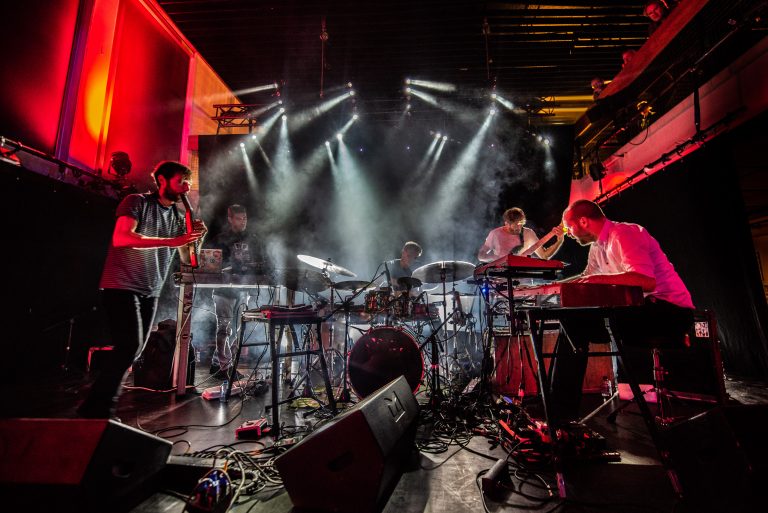 Een muzikale groep speelt op een podium omringd door sfeervolle verlichting en rookeffecten.