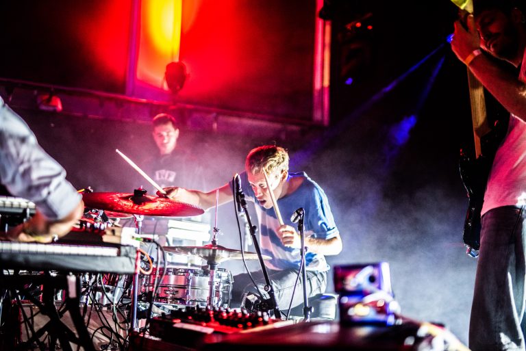 Een drummer speelt energiek op zijn drums tijdens een live concert, met een andere muzikant op de achtergrond.