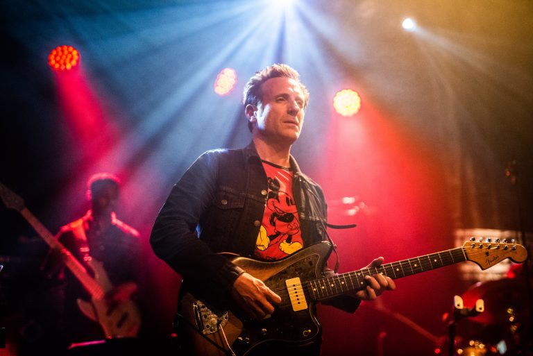 Een muzikant speelt gitaar op een podium met kleurrijke lichtstralen achter zich.