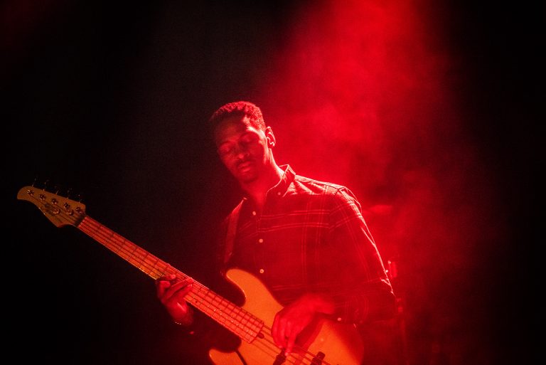 Een muzikant met een elektrische basgitaar staat in een mistige, rode belichting.
