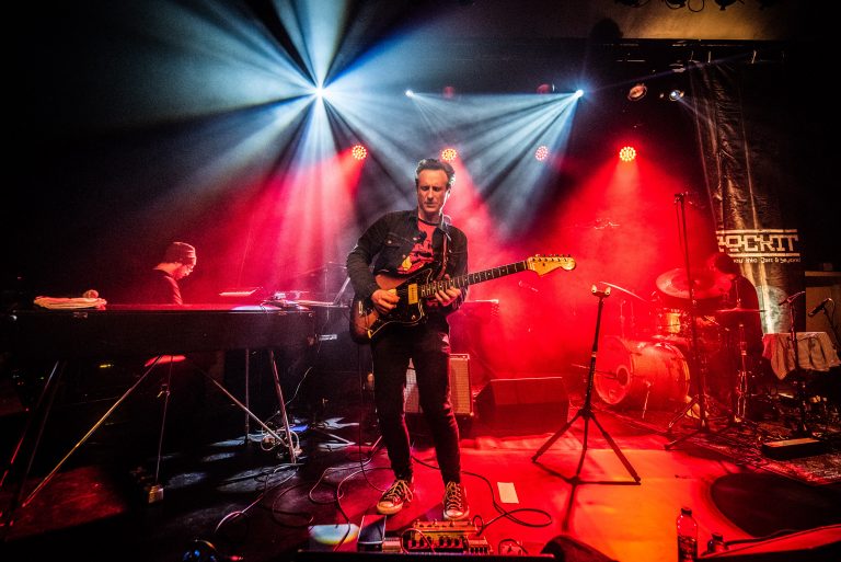Een muzikant speelt gitaar op een podium, omringd door kleurrijke lichtstralen en een band.