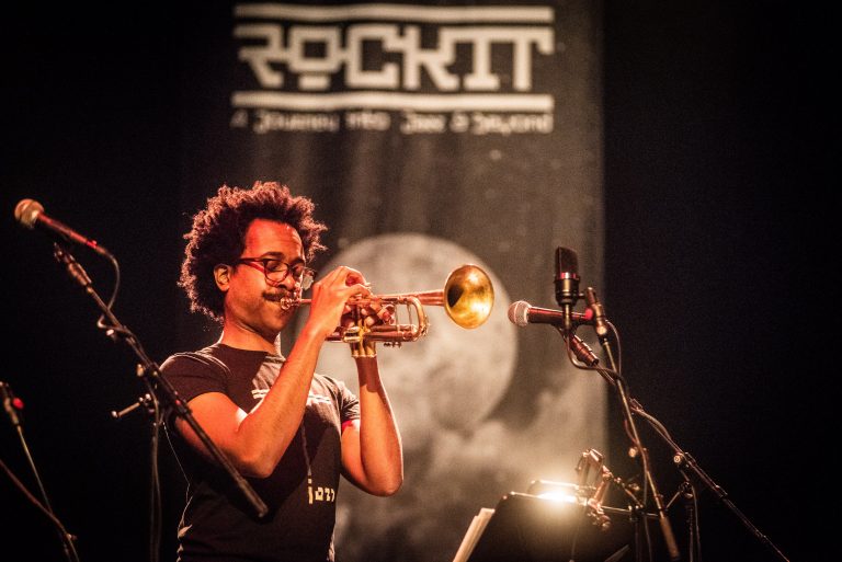 Een muzikant speelt trompet op een podium met een spandoek op de achtergrond.