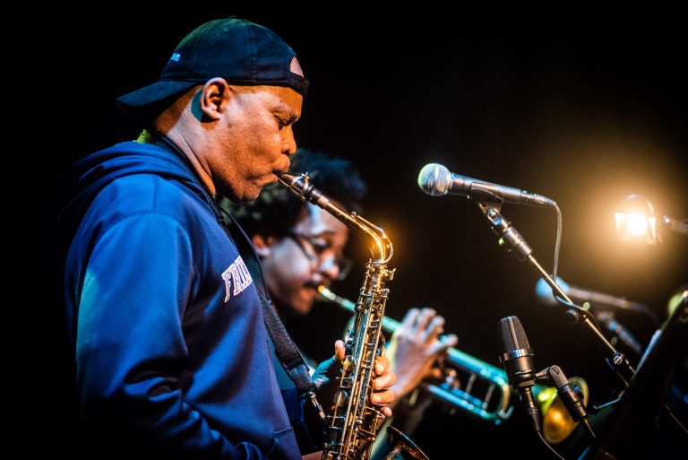 Twee muzikanten spelen op een podium; een man speelt saxofoon terwijl een vrouw op trompet speelt.