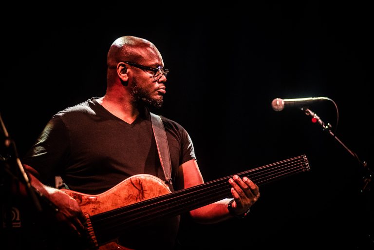 Een muzikant speelt op het podium een elektrische basgitaar onder een donkere achtergrond.