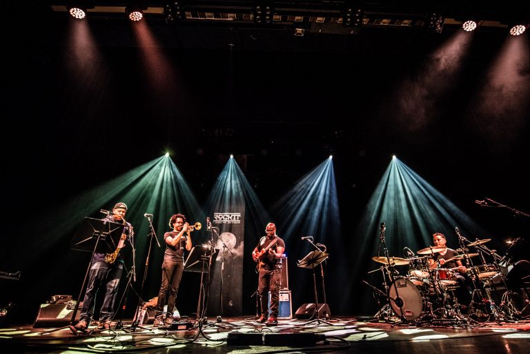Een muzikale bandoptreden met vier leden op het podium, omringd door kleurrijke verlichting.
