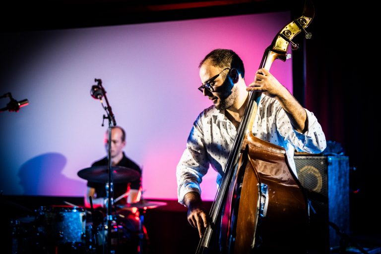 Een muzikant speelt contrabas terwijl een drummer op de achtergrond speelt, omgeven door sfeervolle verlichting.