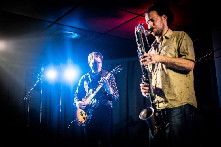 Twee muzikanten optreden op het podium, één met een gitaar en de ander met een saxofoon, verlicht door kleurrijke lichten.