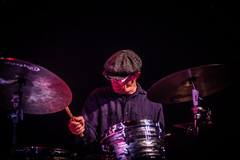 Een drummer speelt op een set met een hoge hoed, gefocust op zijn instrument in een donkere omgeving.