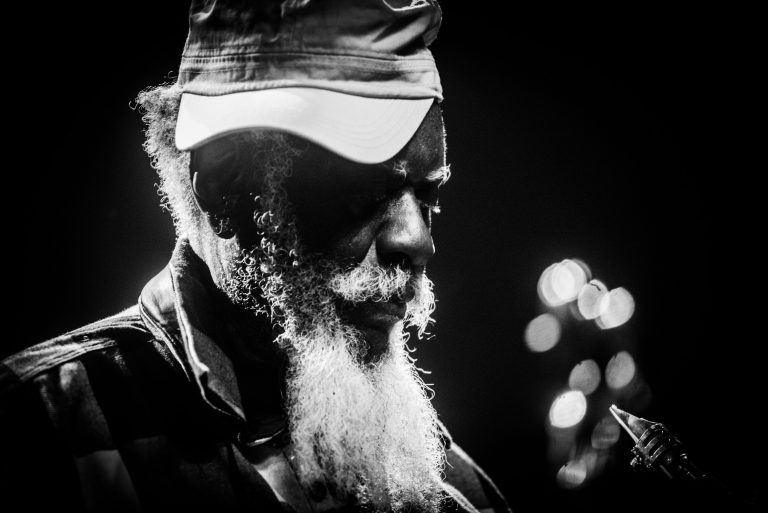 Een oudere man met een baard en een hoed, gefocust op het spelen van een gitaar, verlicht door zachte lichten op de achtergrond.