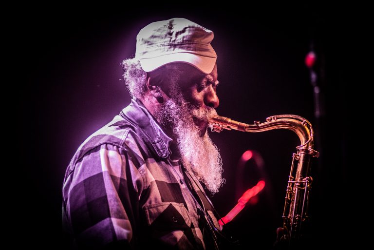 Een muzikant met een baard speelt saxofon, verlicht door kleurrijke lichten.