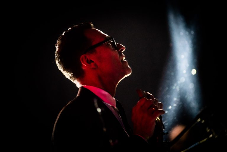 Een man in een zwart kostuum en zonnebril, staande in een spotlicht met een roodachtige gloed, terwijl hij zijn handen in gebed houdt.