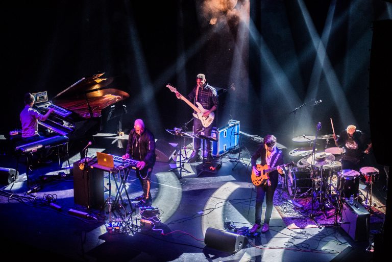 Een live band speelt op het podium met verschillende instrumenten en kleurrijke verlichting.