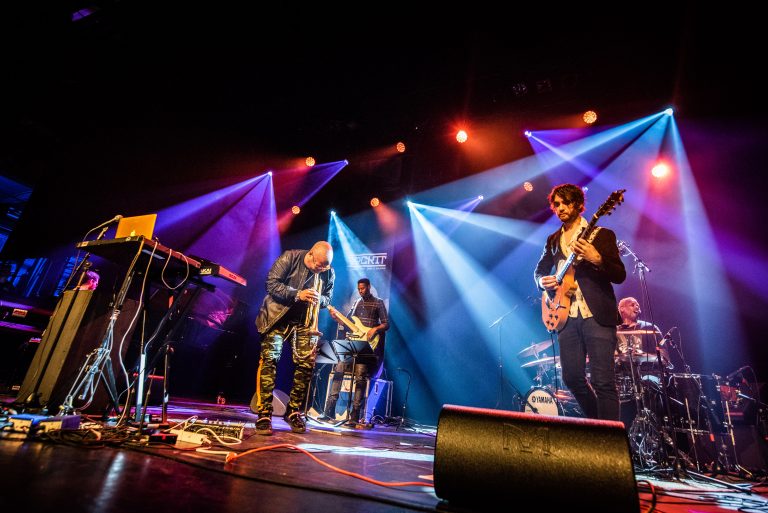 Een band performt op een verlichte podium met meerdere muzikanten die verschillende instrumenten bespelen.