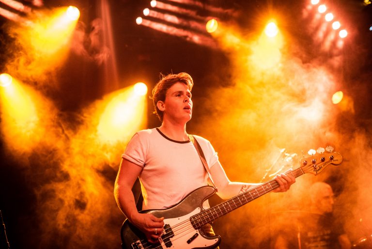 Een muzikant speelt basgitaar op een podium, omgeven door kleurrijke verlichting en lichteffecten.