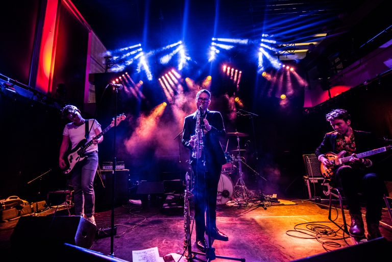 Een band speelt live op een podium, omgeven door kleurrijke lichten en rook effecten.