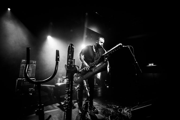 Een muzikant staat op een podium met een groot muziekinstrument, verlicht door dramatische lichtstralen.