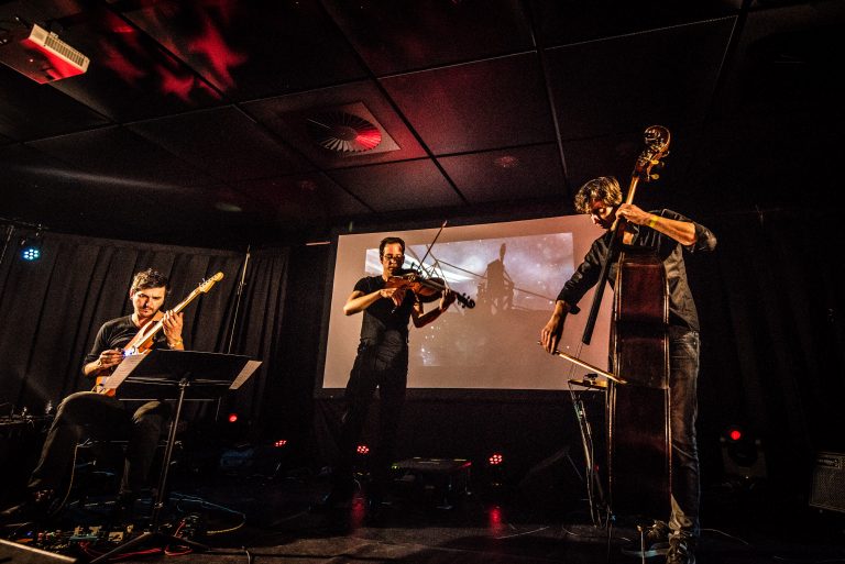 Drie muzikanten spelen op een podium met een projectiescherm op de achtergrond.