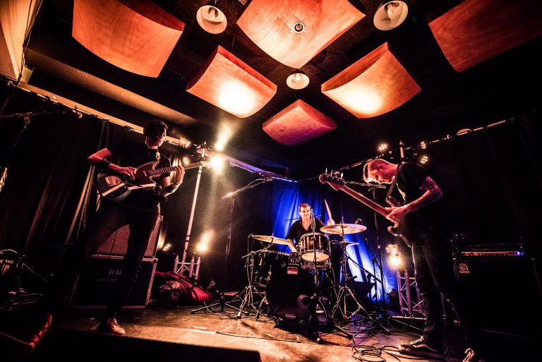 Een band speelt live op een podium met muzikanten die gitaar en drums bespelen, omringd door sfeervolle verlichting.