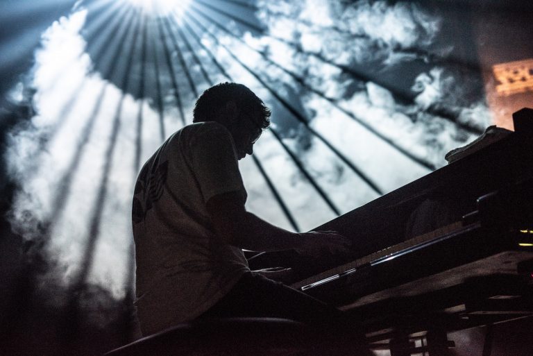 Een silhouet van een persoon die een piano speelt, met stralen van licht achter hem in een bewolkte lucht.