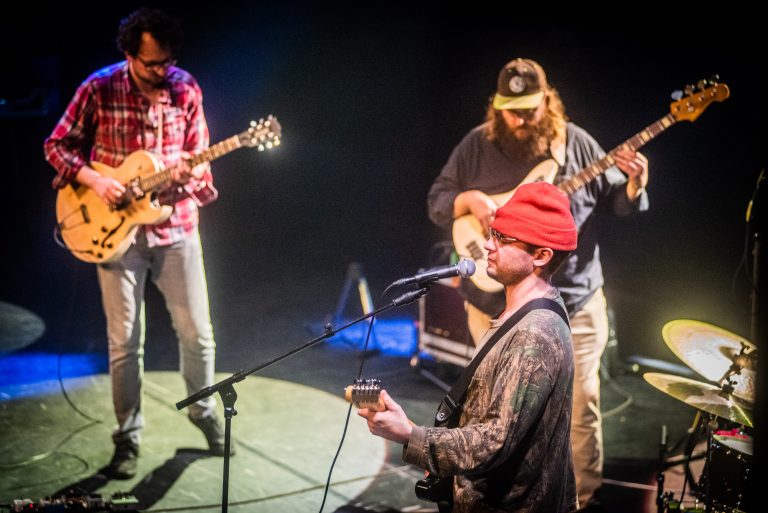Drie muzikanten optreden op het podium met verschillende gitaren en een microfoon.