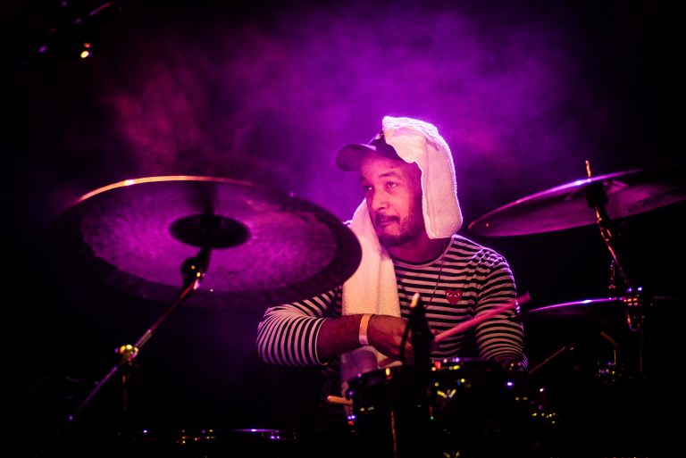 Een drummer in een gestreept shirt, met een handdoek op het hoofd, speelt op een set met paarse belichting.