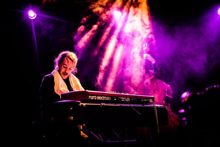 Een muzikant zit achter een keyboard, omringd door paarse lichten en rook.