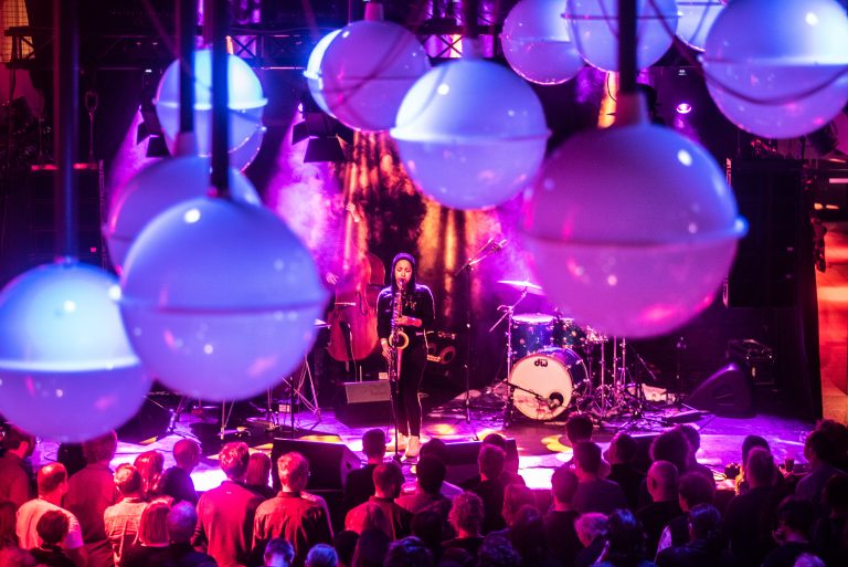 Een live optreden in een donker verlichte zaal met kleurrijke verlichting en grote, ronde lampen aan het plafond.
