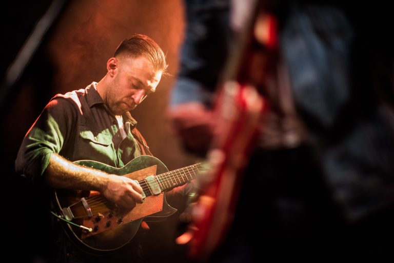 Een muzikant speelt gitaar op het podium, gefocust op zijn instrument terwijl een andere gitaar op de voorgrond zichtbaar is.