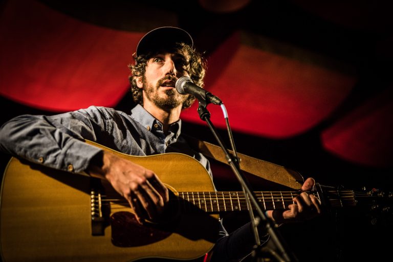 Een muzikant speelt gitaar en zingt in een intieme podiumsetting met rode verlichting.