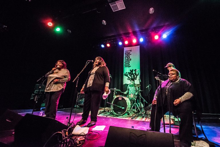 Een groep zangers staat op een podium met microfoons, omringd door kleurrijke verlichting en een drumset op de achtergrond.