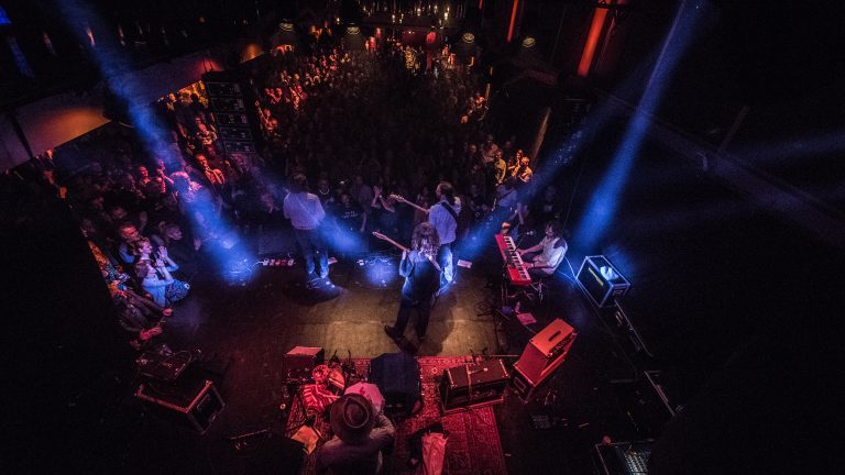 Een band speelt live op een podium voor een groot publiek, verlicht door kleurrijke lichten.